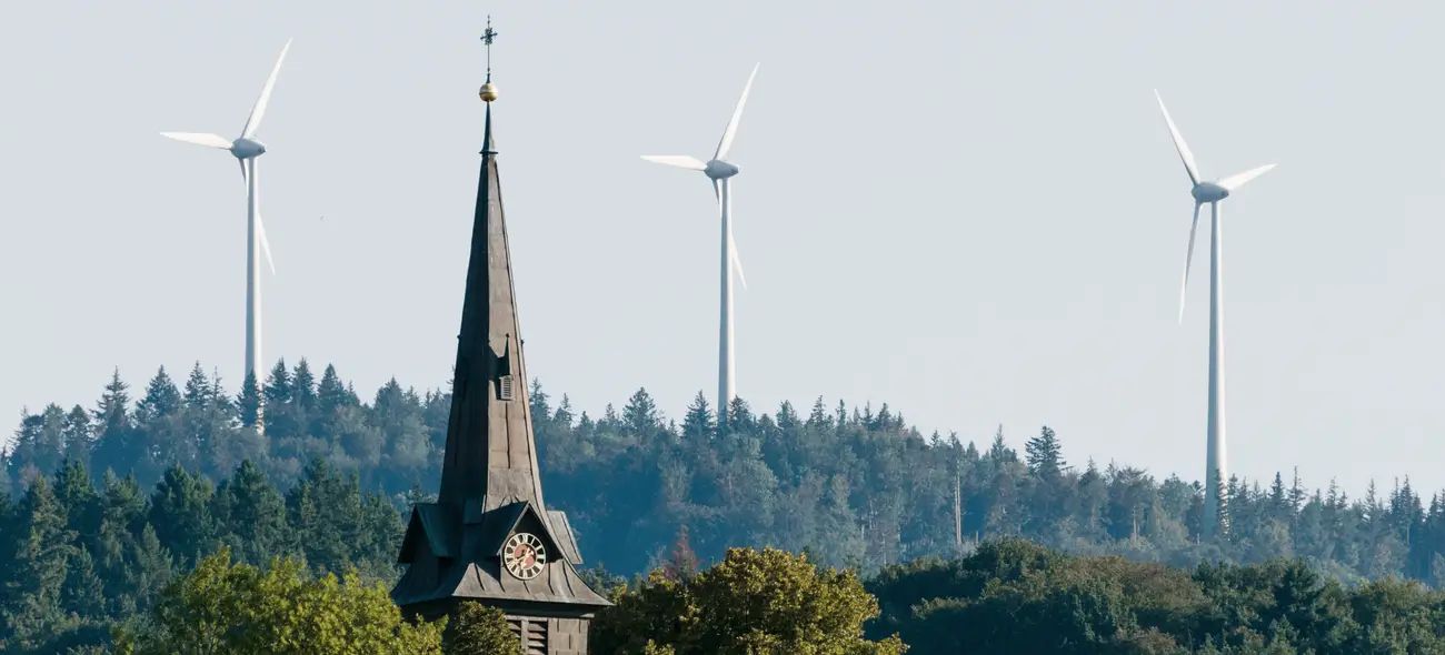 Dorf mit Kirchturm und Windkraftanlagen im Hintergrund.