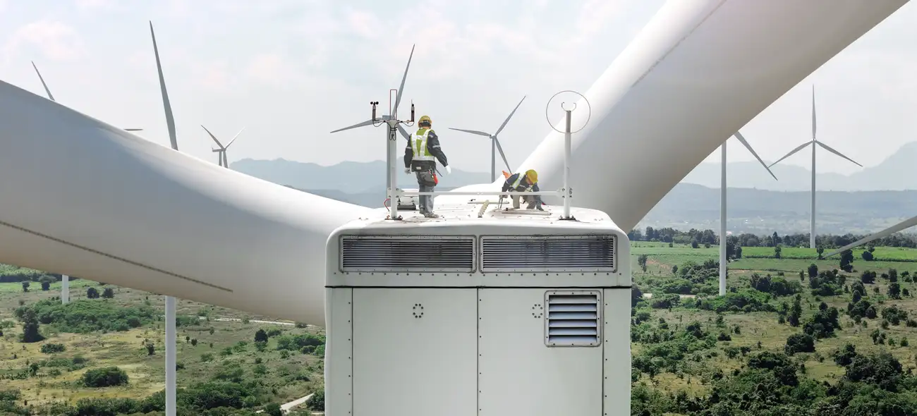 Techniker stehen auf einer Windkraftanlage und inspizieren einen weiteren Windpark in hügeliger Landschaft.