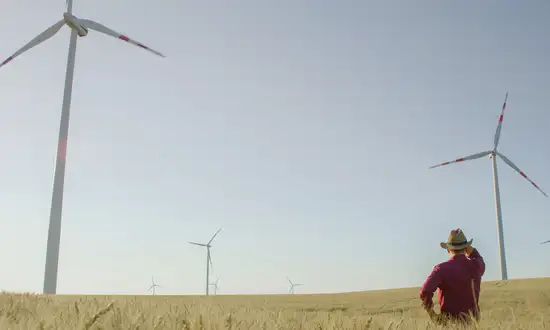 Welche Pachtpreise Grundeigentümer mit Windrädern erzielen Person mit rotem Hemd und Hut steht auf einem Feld und blickt auf Windräder in der Nähe.