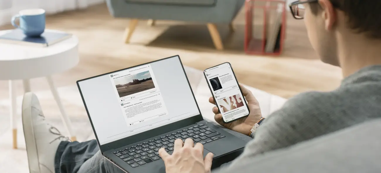 Person sitzt, von hinten gesehen, mit Laptop auf dem Schoß, hält Smartphone, beide zeigen Webseiten an.