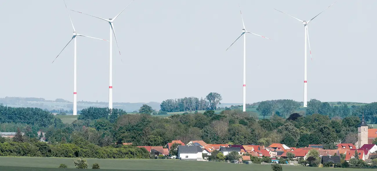 Windenergie in Kommunen – wie Städte und Gemeinden die Energiewende zum Heimspiel machen Mehrere Windräder stehen hinter einem Dorf mit roten Dächern und grünen Bäumen in ländlicher Landschaft.