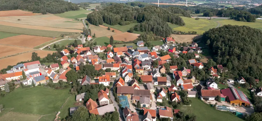 Luftaufnahme eines Dorfes mit mehreren Windrädern in hügeliger Landschaft im Hintergrund.