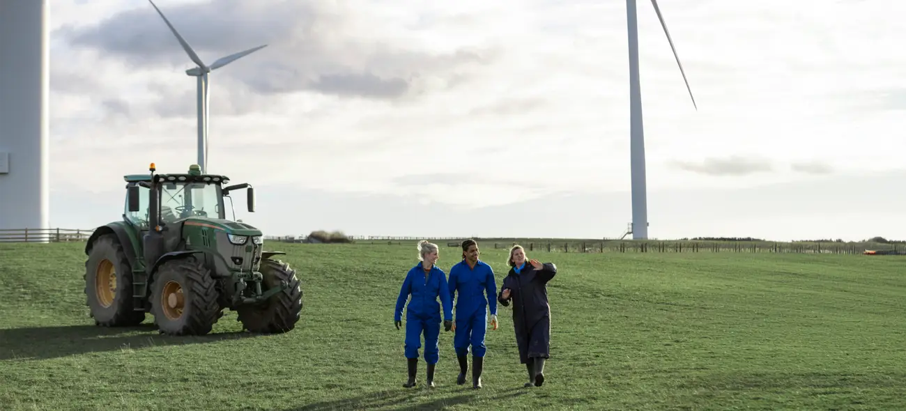 Windenergie in der Landwirtschaft durch Flächenverpachtung Drei Landwirte auf einem Feld mit einem Traktor und Windrädern im Hintergrund.